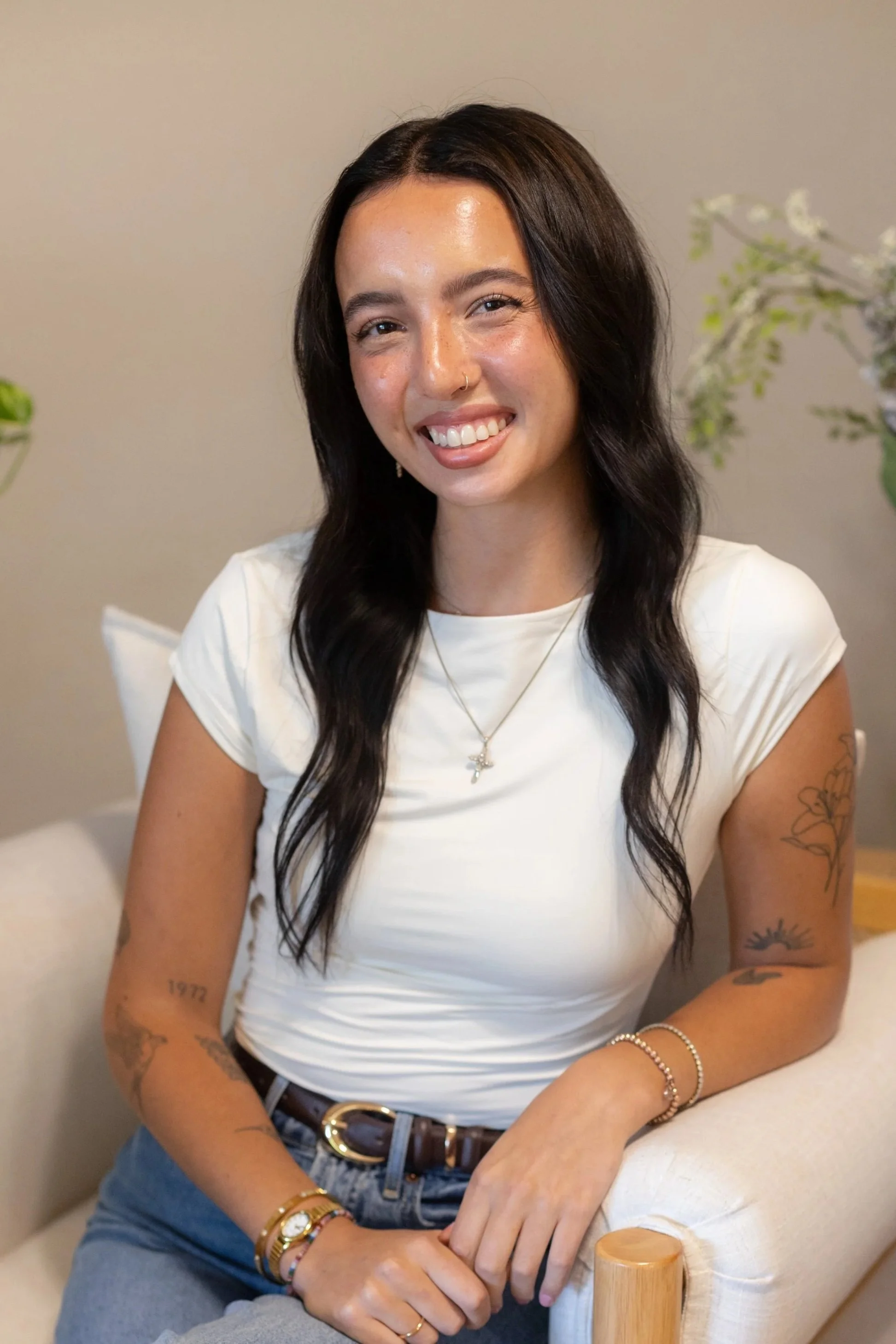 A young woman with dark wavy hair, tattoos on her arms and a nose ring, smiling and sitting on a beige armchair with a wooden armrest, wearing a white t-shirt and jewelry.