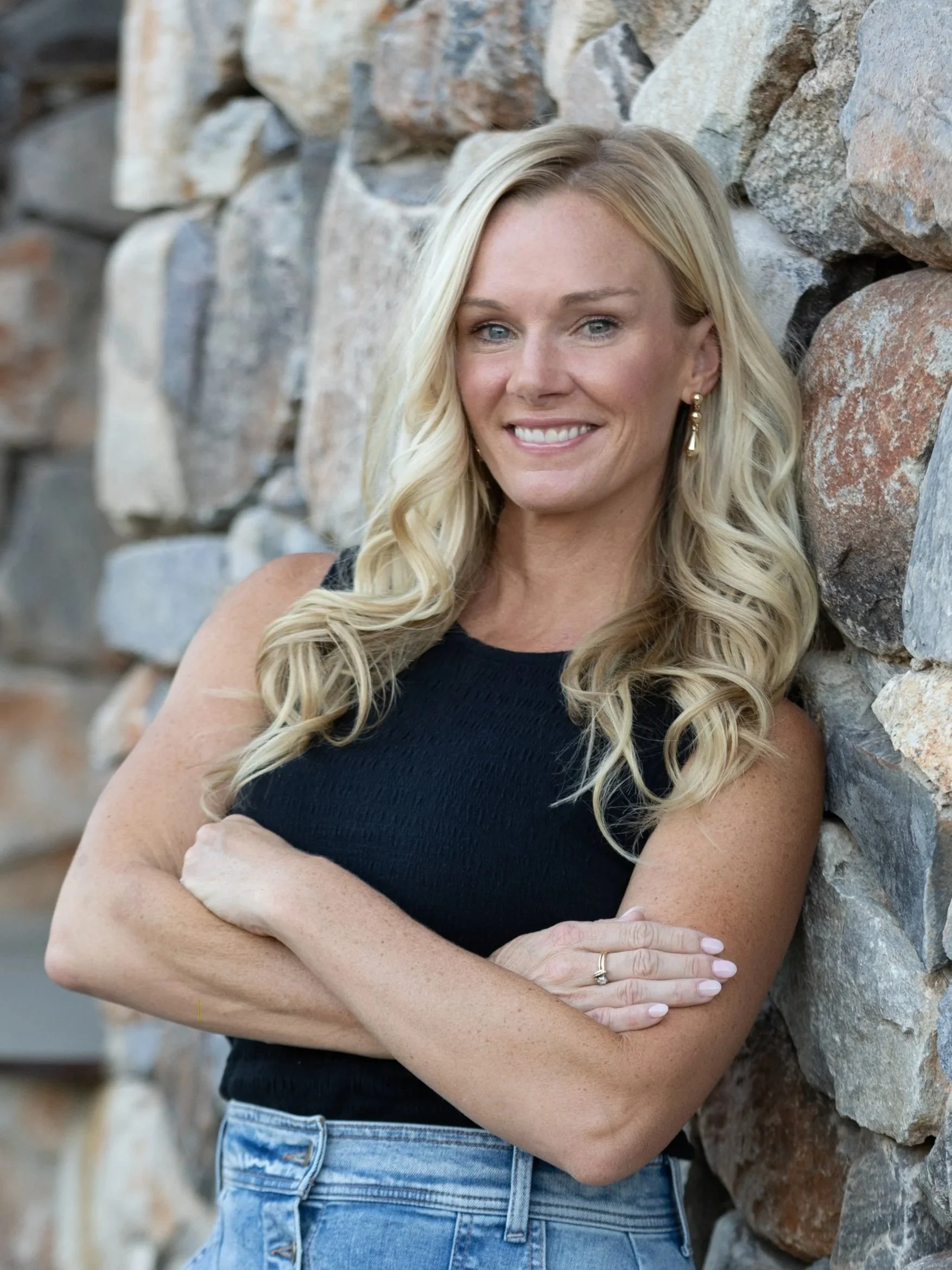 A woman with long blonde hair, wearing a black sleeveless top and blue jeans, standing with arms crossed in front of a rock wall.