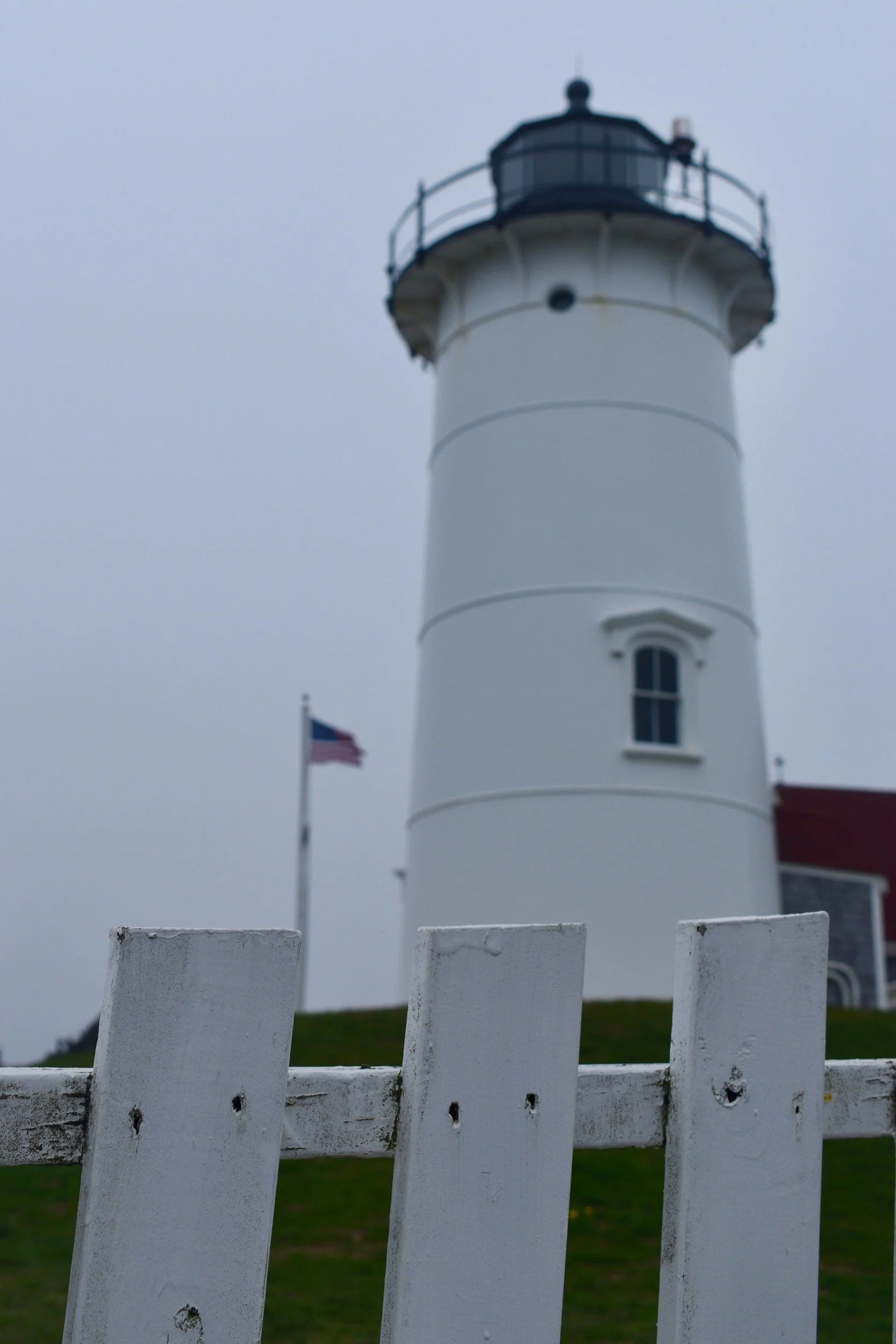 A white lighthouse with a black lantern room at the top, behind a white picket fence and an American flag on a flagpole, on a cloudy day.