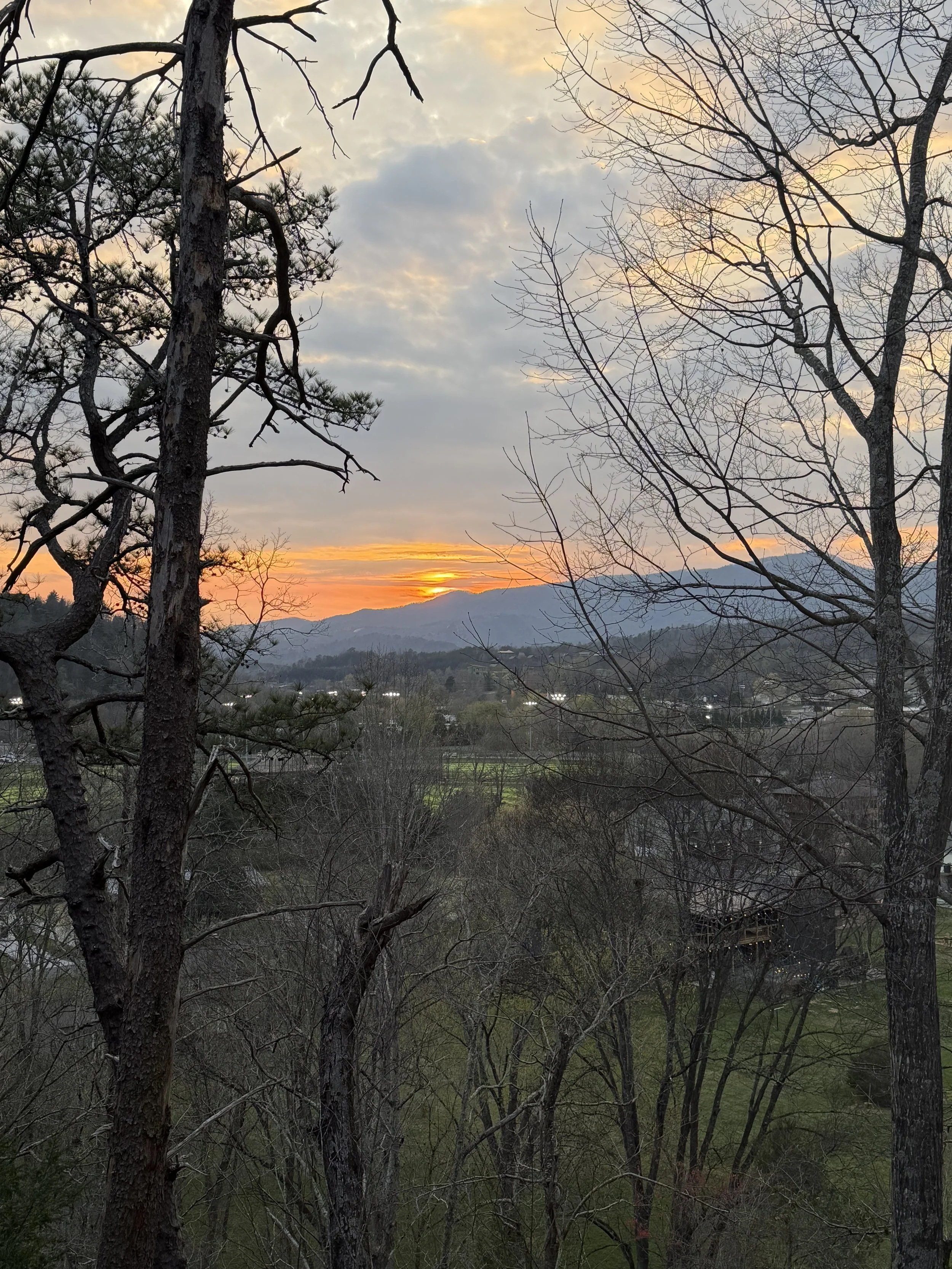 A scenic landscape at sunset with a sky partly cloudy, distant mountains, and trees in the foreground.
