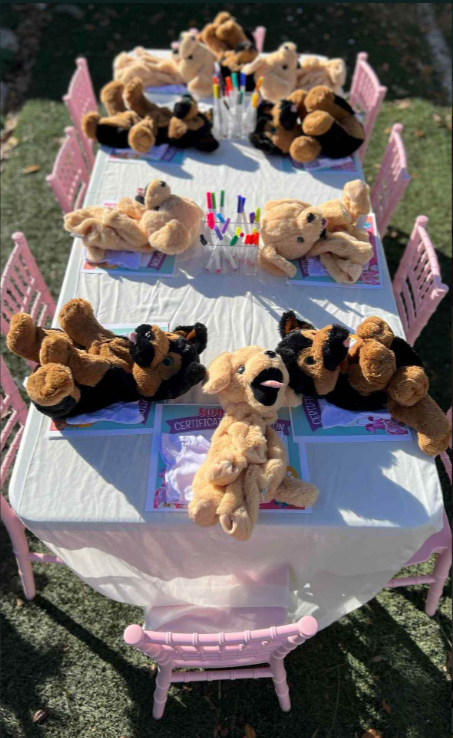 A birthday party table decorated with stuffed animal puppies and pink chairs outside.