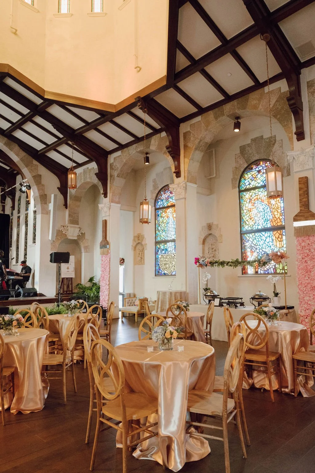 Elegant indoor event space decorated for a celebration with round tables covered in blush-colored satin tablecloths, floral centerpieces, stained glass windows, and a stage with musicians.