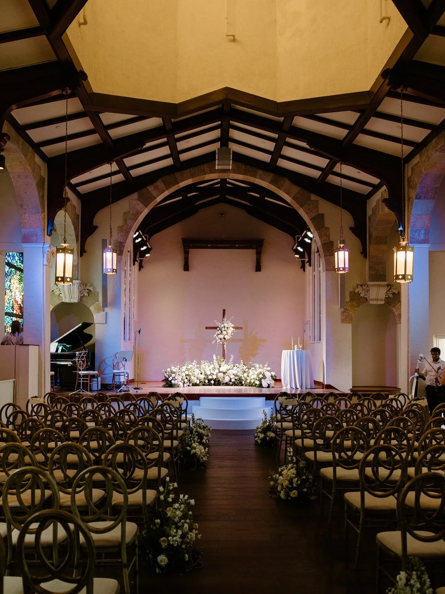 Empty indoor wedding chapel with wooden chairs decorated with flowers along the aisle, a floral altar with a cross, and a grand piano to the left.