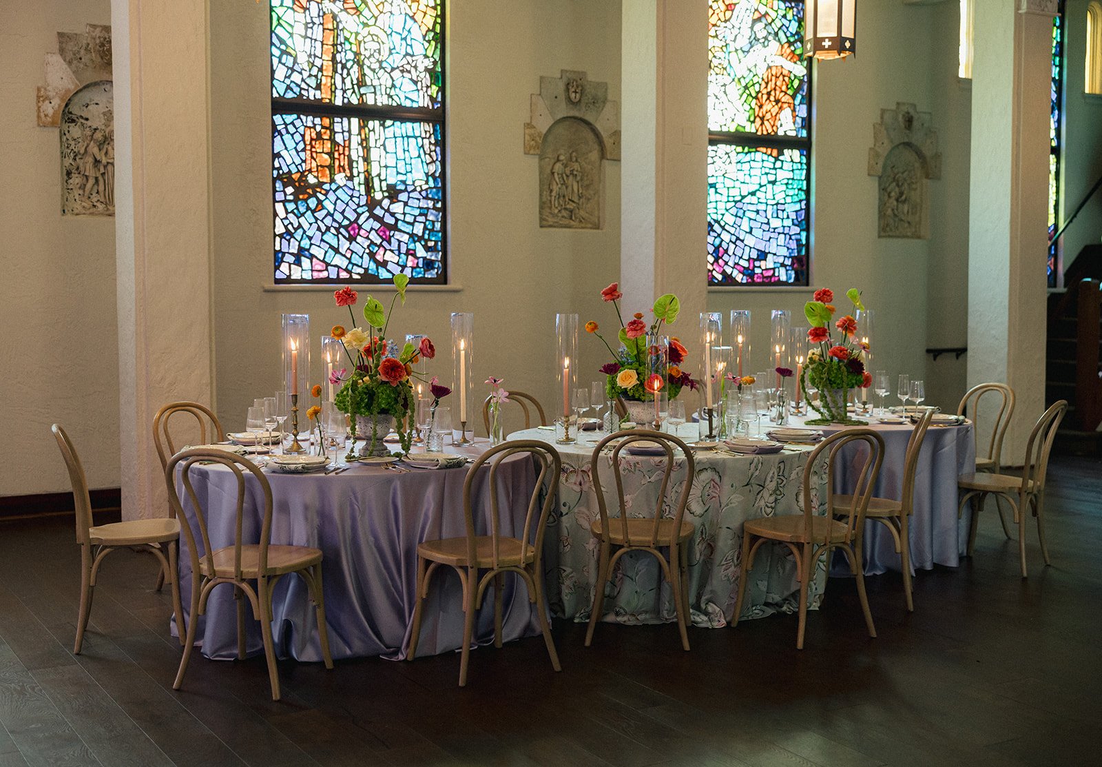 A round table set for a formal event with floral centerpieces, candles, and glassware in a room with stained glass windows at Gather Lake Wales.