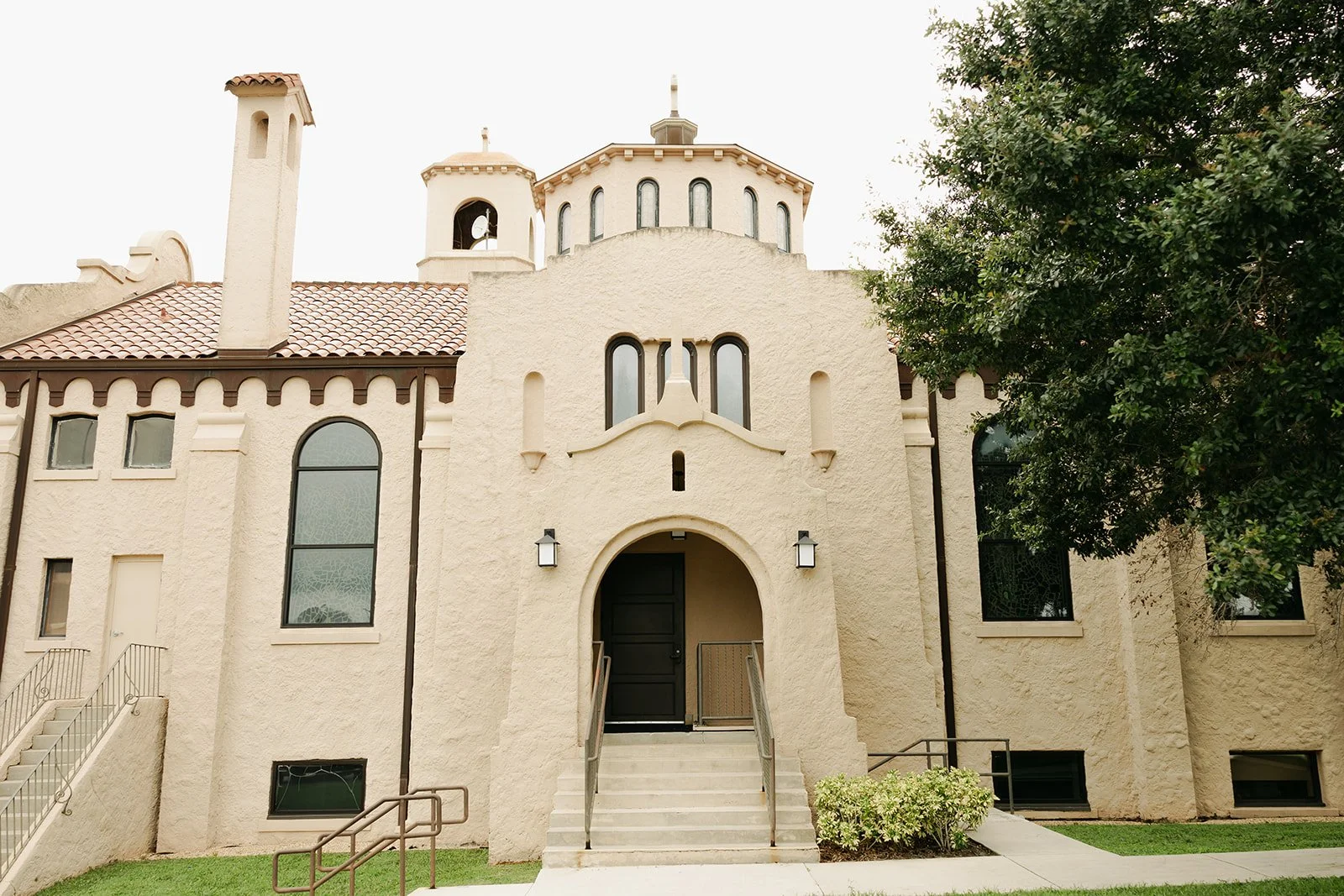 Gather at Lake Wales Arts Center, A beige stucco building with arched windows and a black door, set in a landscaped area with a pathway, stairs, and shrubs, and a tree on the right side.
