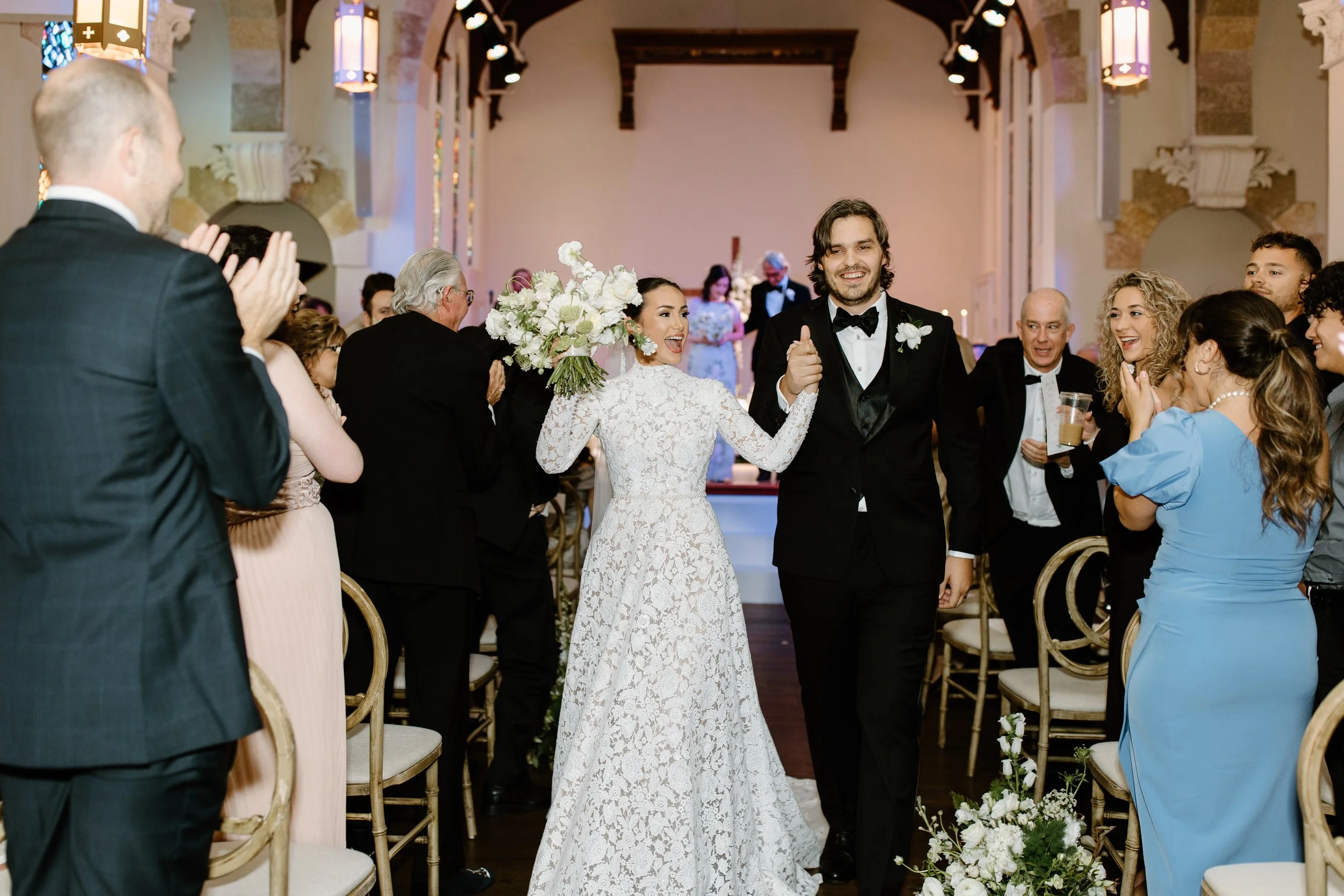 A newlywed couple walking down the aisle at Gather at Lake Wales Art Center smiling and holding hands, surrounded by clapping and happy guests in an elegant venue.