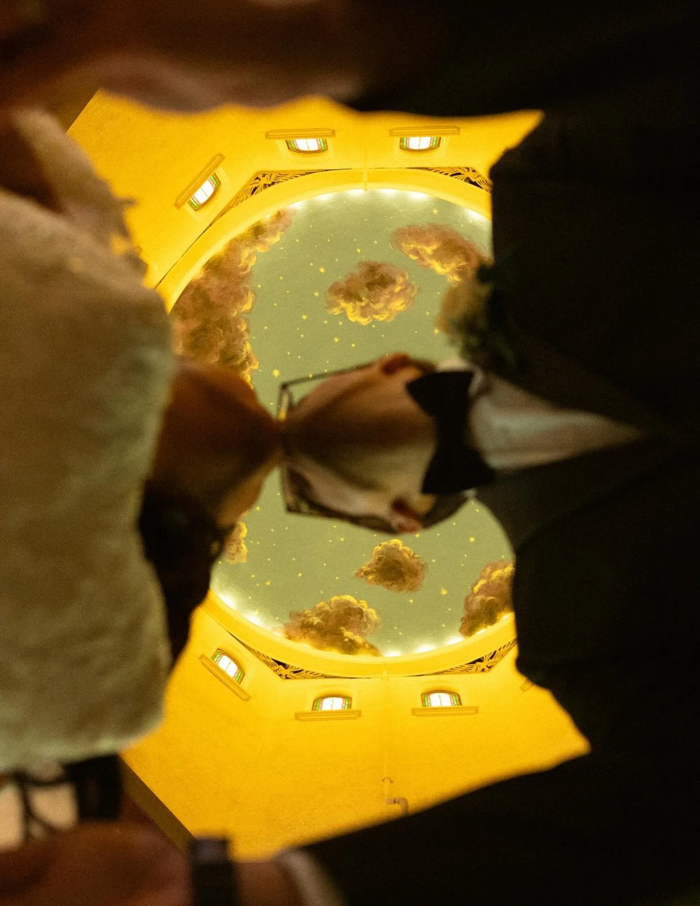 A person dressed in formal attire, including a tuxedo with a bow tie and glasses, taking a photo from the ground with a decorated ceiling featuring clouds, stars, and a yellow border.