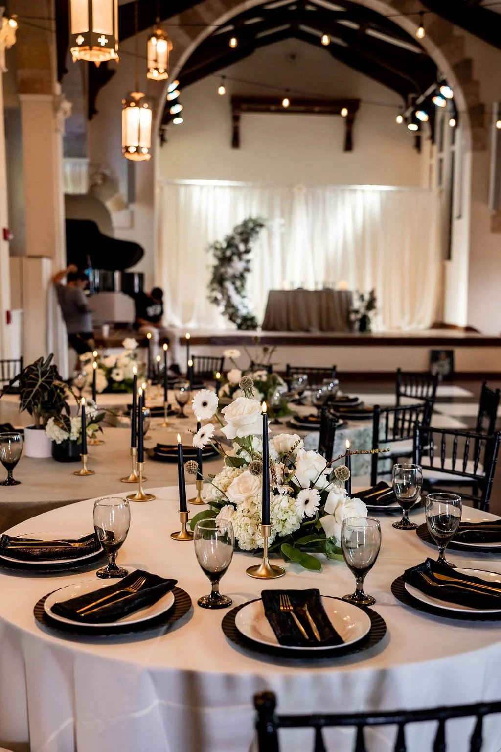 Elegant banquet table set with black and white dinnerware, glassware, and black candles on gold candleholders, decorated with white floral centerpieces and greenery, in a decorated event space with draped curtains and a stage in the background.