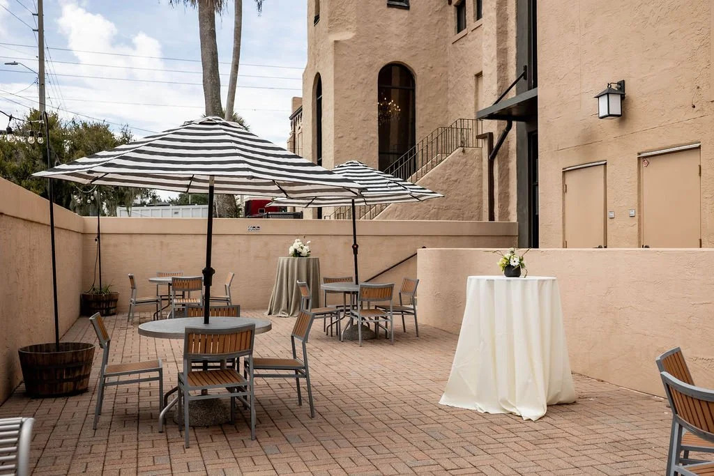 An outdoor patio with round tables, some with umbrellas and others with tablecloths, and chairs, decorated with flowers in vases, against a beige stucco building.