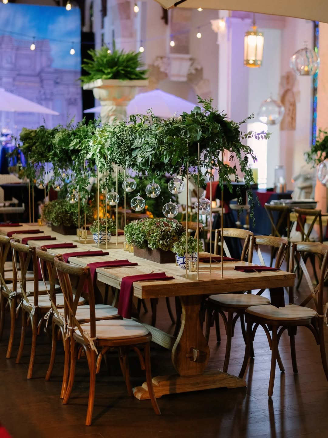 An elegant event setup with a wooden table decorated with green foliage and hanging glass globes, surrounded by wooden chairs with red napkins, in a warmly lit indoor space.