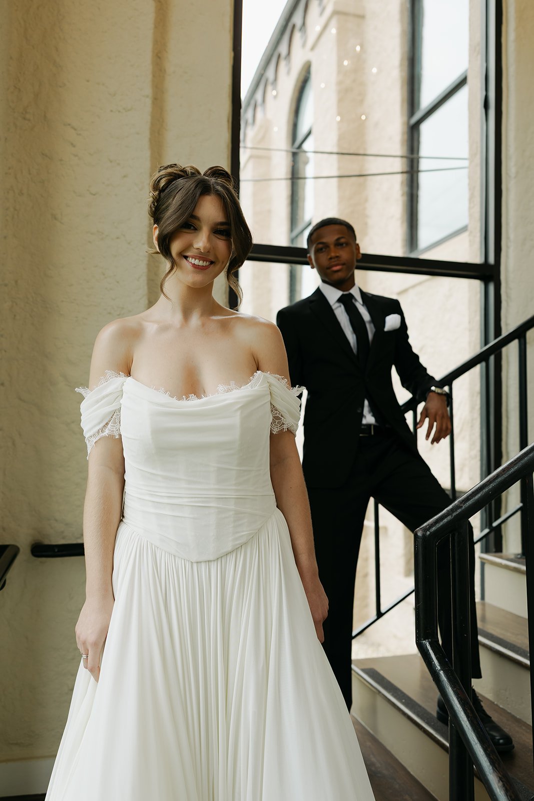 A smiling woman wearing a white off-shoulder wedding dress standing at the bottom of a staircase, with a man in a black suit and tie leaning on the railing in the background.