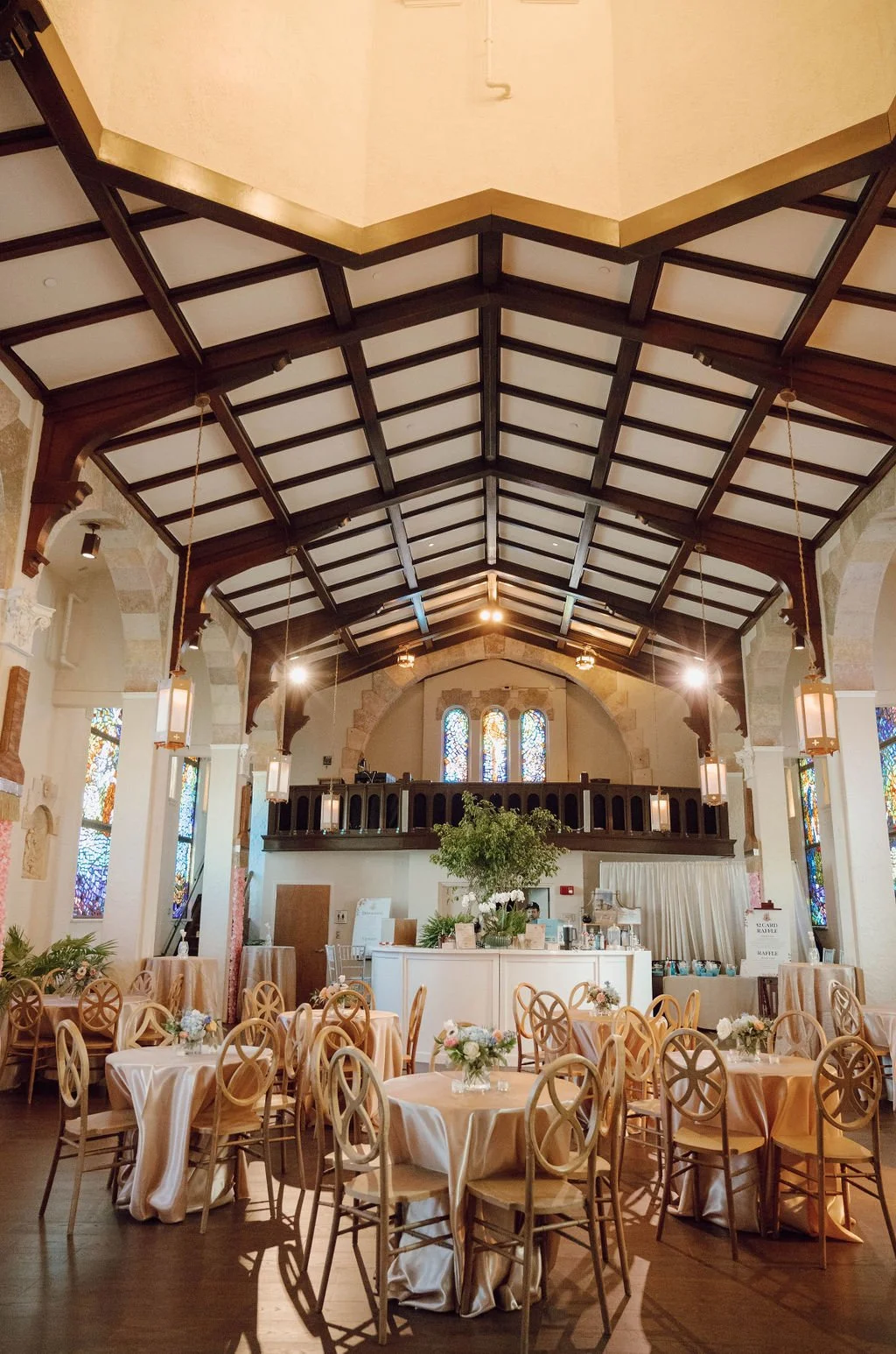 Elegant dining hall with high vaulted ceiling, stained glass windows, and round tables covered with beige tablecloths and flower arrangements.