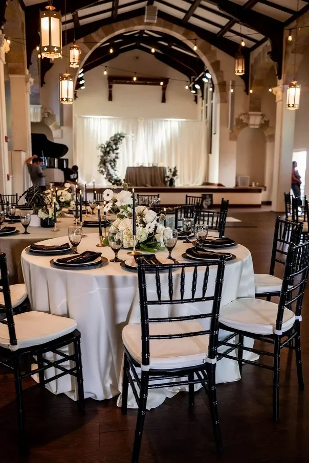 Elegant banquet hall with a round table decorated with white tablecloth, black napkins, black chairs with white cushions, and floral centerpieces, set up for an event or wedding reception. In the background, there's a stage with a white curtain backd