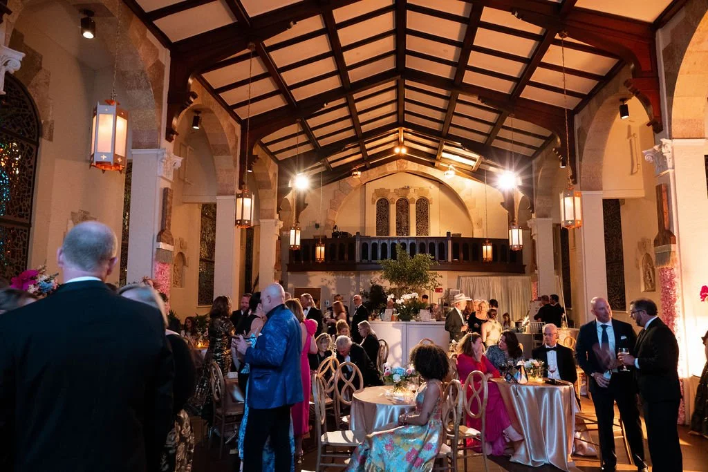 Guests mingling at a formal event or wedding reception inside a decorated hall with high vaulted ceilings, wooden beams, and ornate windows.
