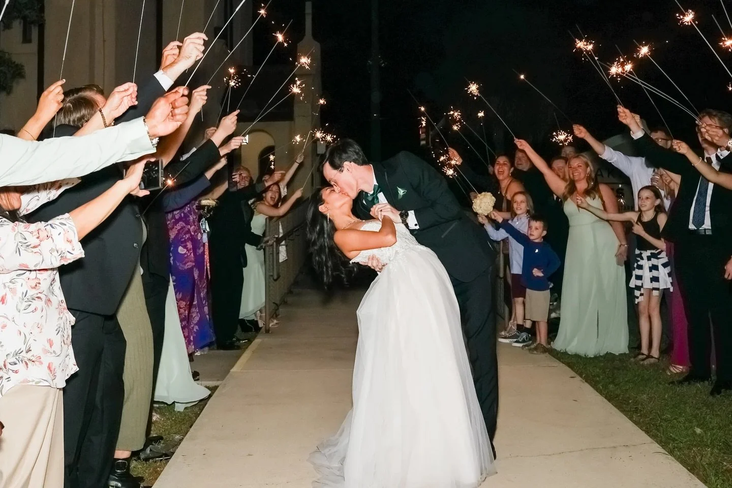 A bride and groom share a kiss under sparklers held by friends and family during a wedding celebration at night.