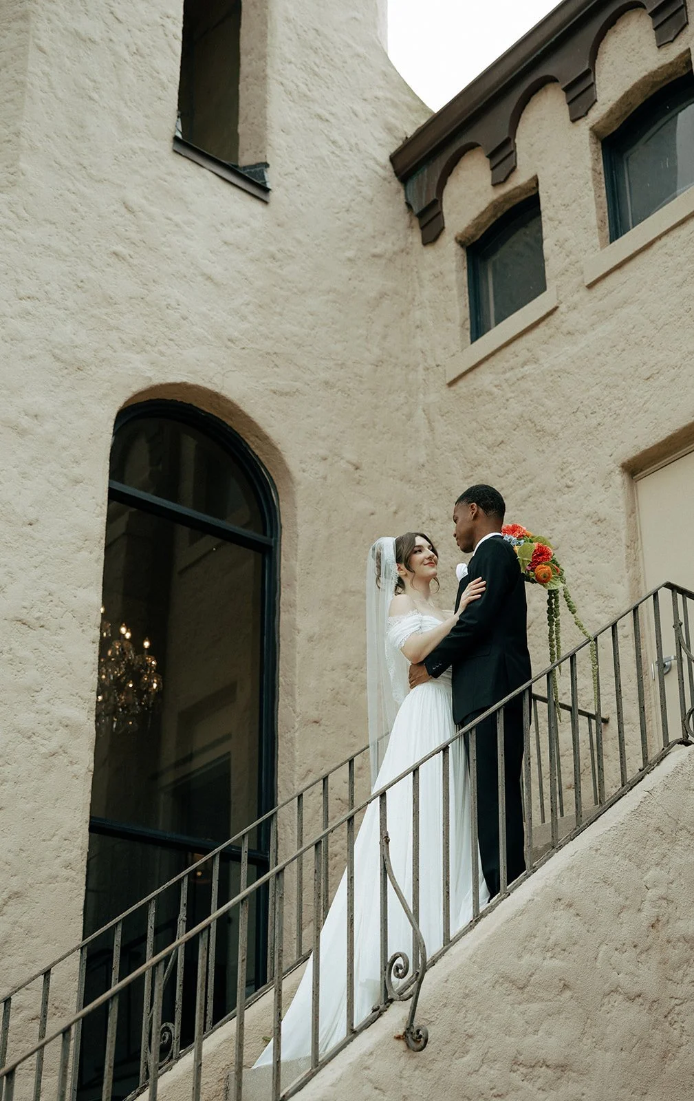 A bride and groom sharing a kiss on a staircase outside a beige stucco building with large arched windows.