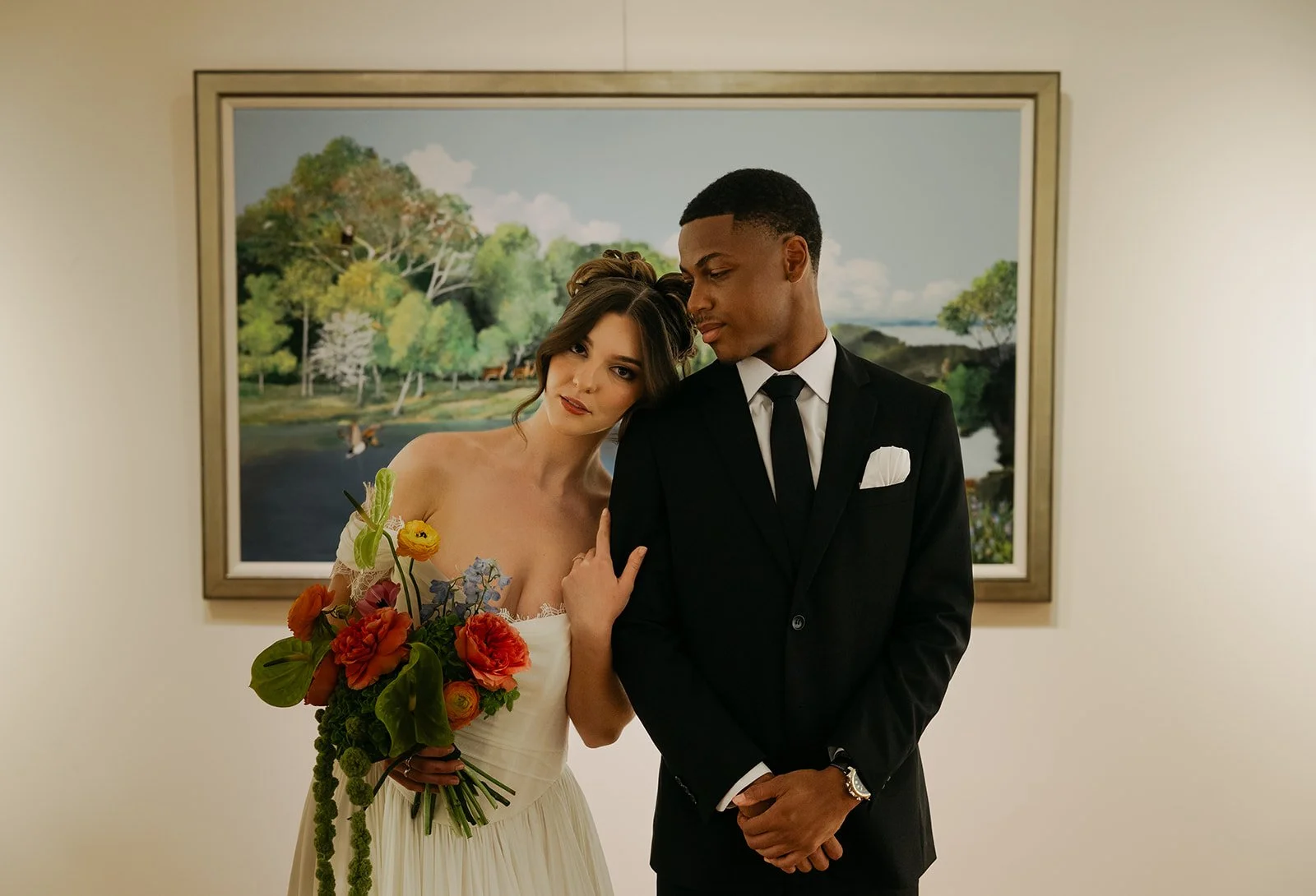A bride and groom standing together at a wedding, with the bride holding a bouquet of colorful flowers, in front of a landscape painting.
