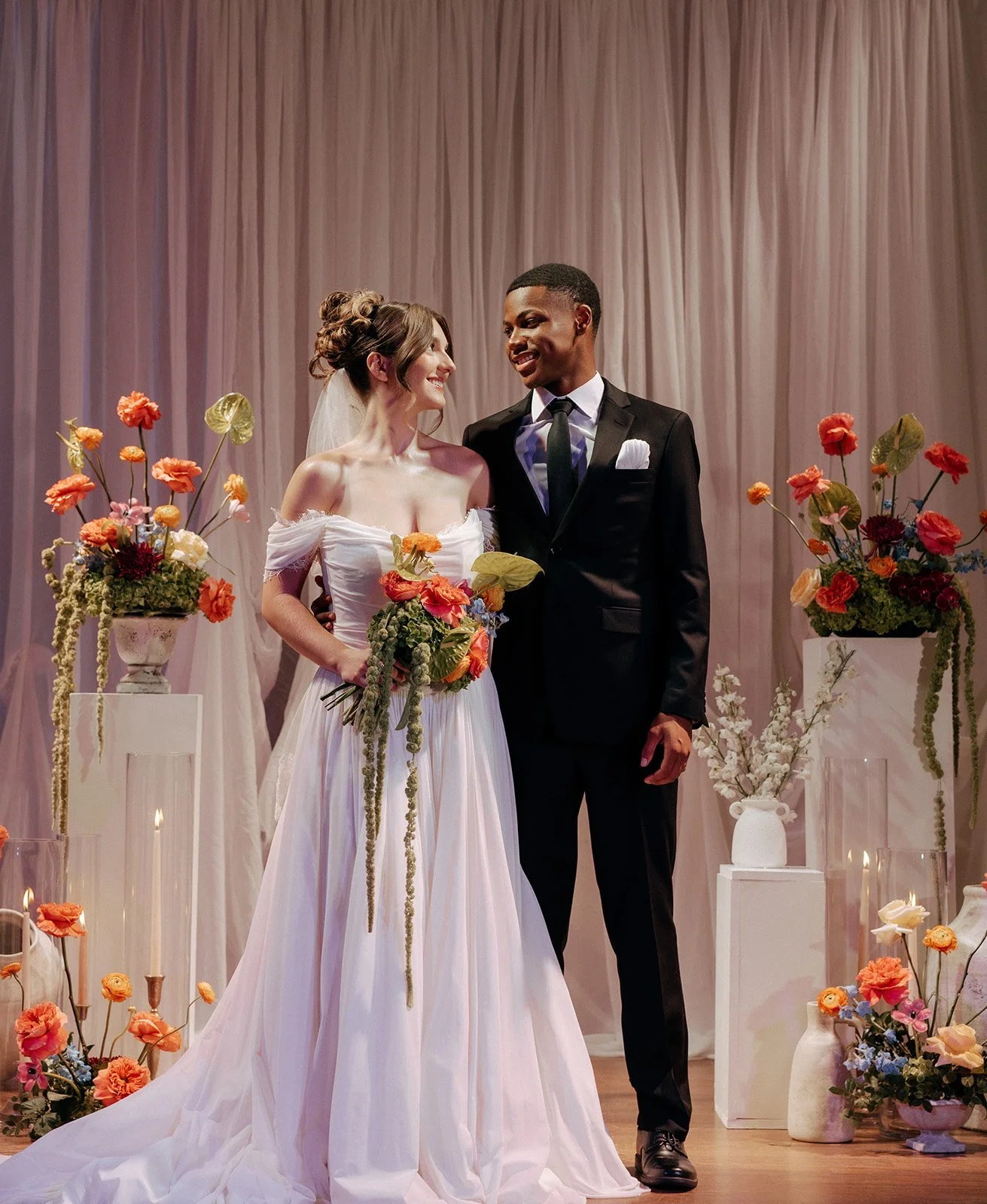 A bride and groom standing together at their wedding ceremony, surrounded by floral arrangements and candles, exchanging looks with a pink curtain backdrop.