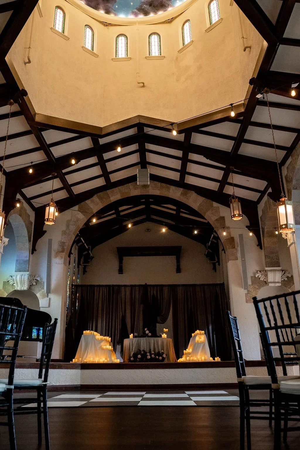 Interior of a decorated venue, possibly for a wedding or special event, with candles on tables and a stage area with dark curtains. The ceiling features a semi-circular opening with stained glass windows and a starry sky mural. Hanging lights are str