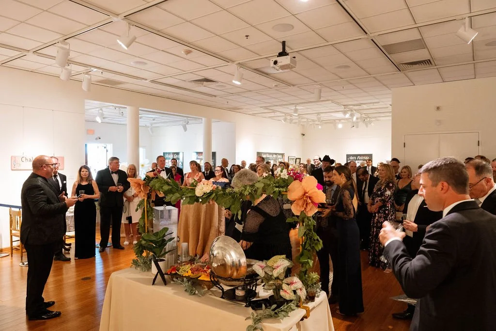 A group of people in formal attire attending a social event or reception inside a gallery or event space, with a table decorated with flowers and ornaments in the foreground.