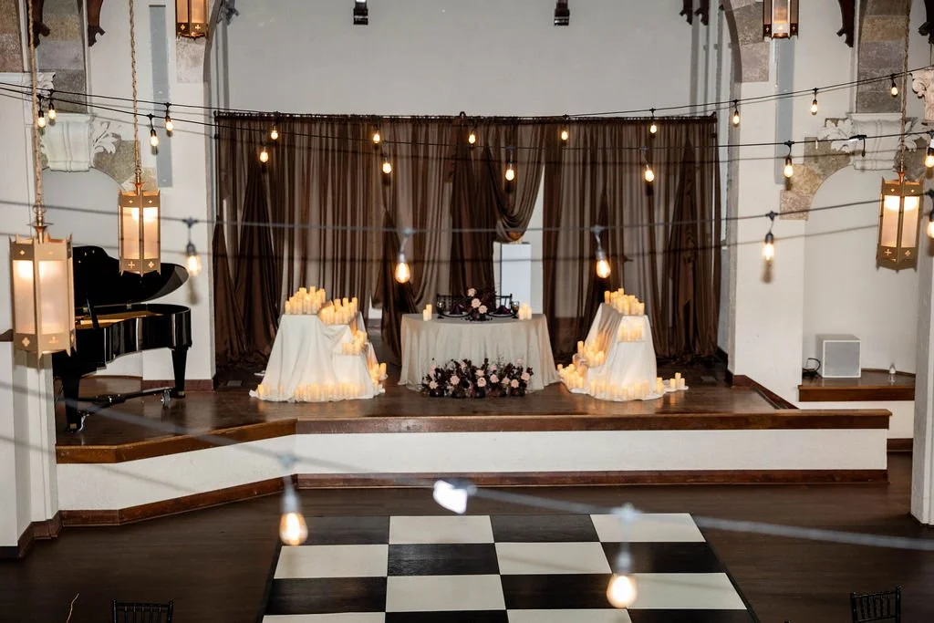 Wedding ceremony stage decorated with candles, flowers, and drapes, with hanging string lights and lanterns in a grand hall with a checkered dance floor.