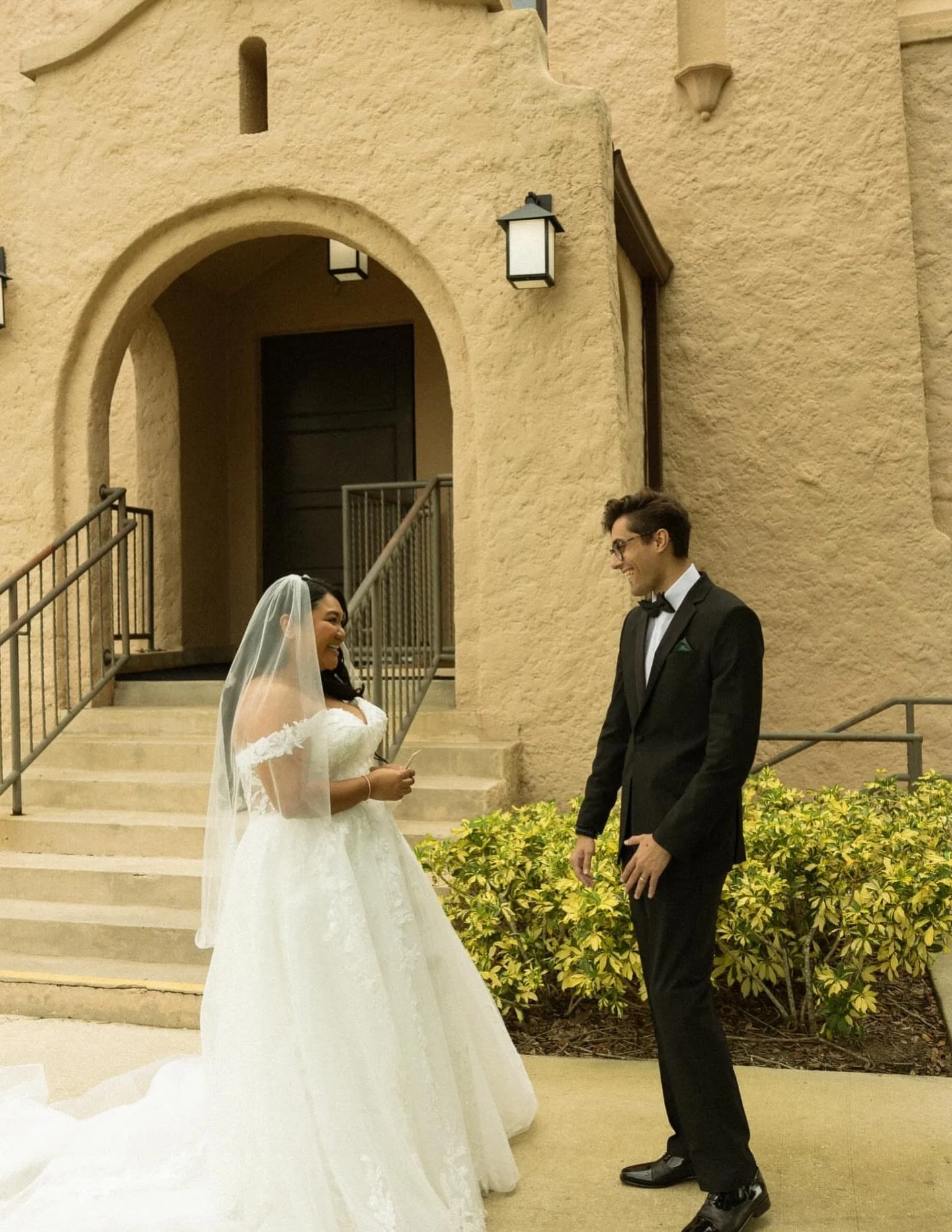 A bride and groom smiling at each other outside a church or similar building. The bride is wearing a white wedding dress and veil, while the groom is in a black tuxedo with a bow tie.