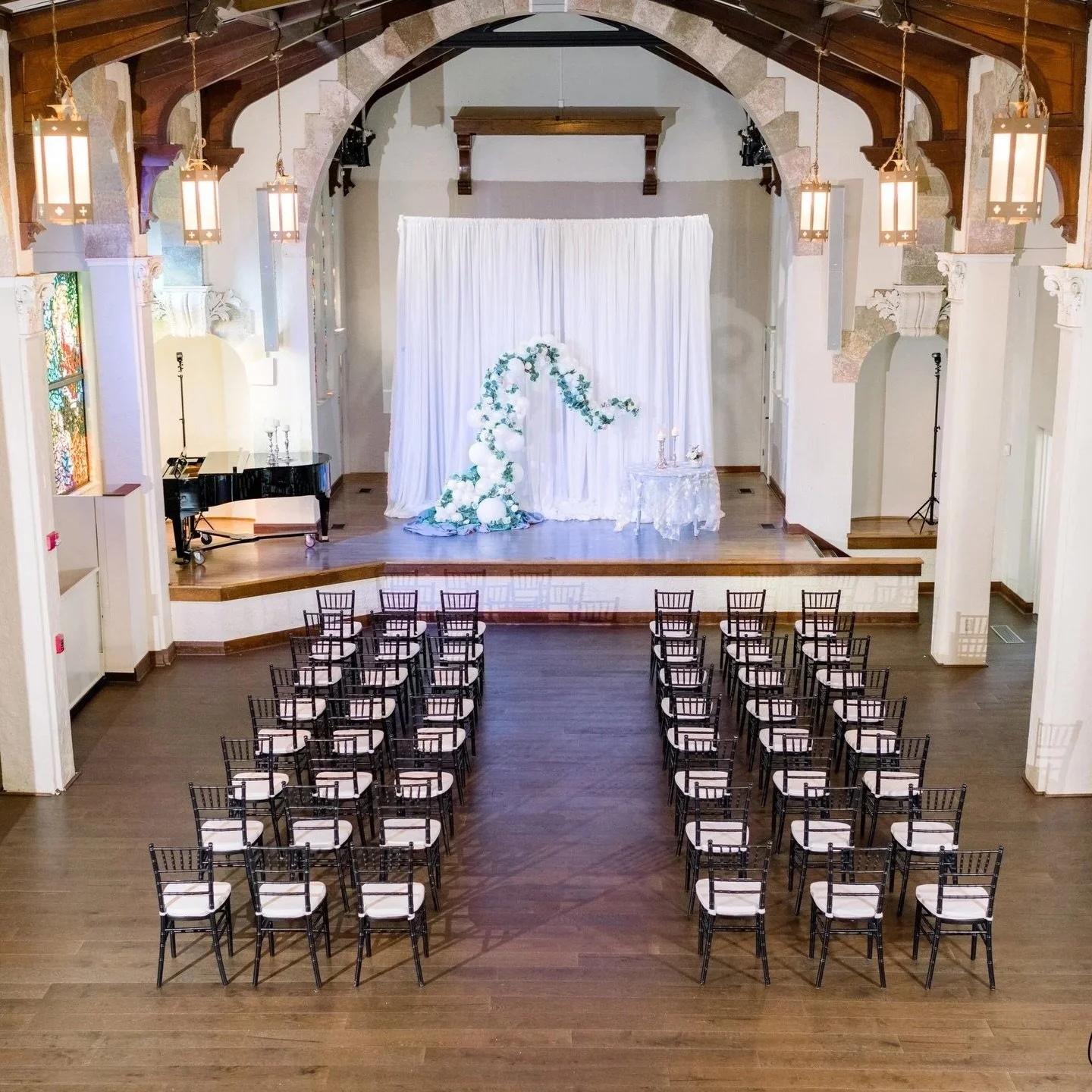 Wedding ceremony setup with chairs facing an elegant stage decorated with white drapery and floral arrangement, pianos, and candles.