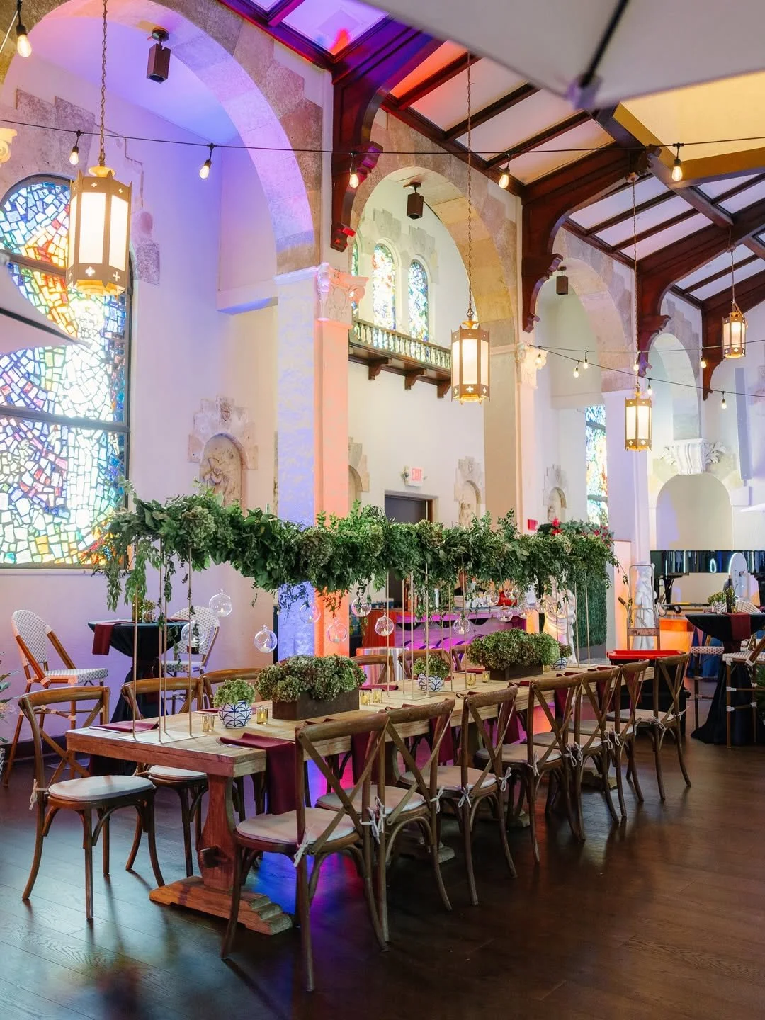 Elegant event space with wooden tables and chairs, adorned with green foliage centerpieces and hanging glass ornaments, set inside a church-like venue with stained glass windows, arched ceiling, and warm lighting.