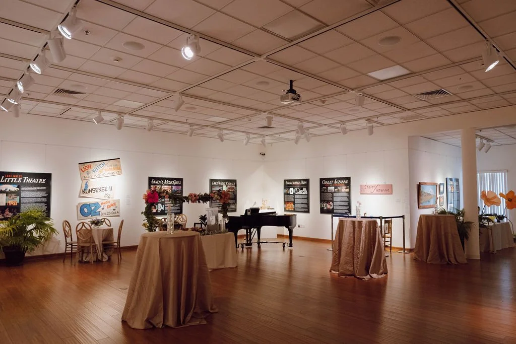 An art gallery with framed posters on the white walls, a black grand piano in the center, and tables with beige tablecloths and floral arrangements, illuminated by ceiling spotlights and natural light from a window.
