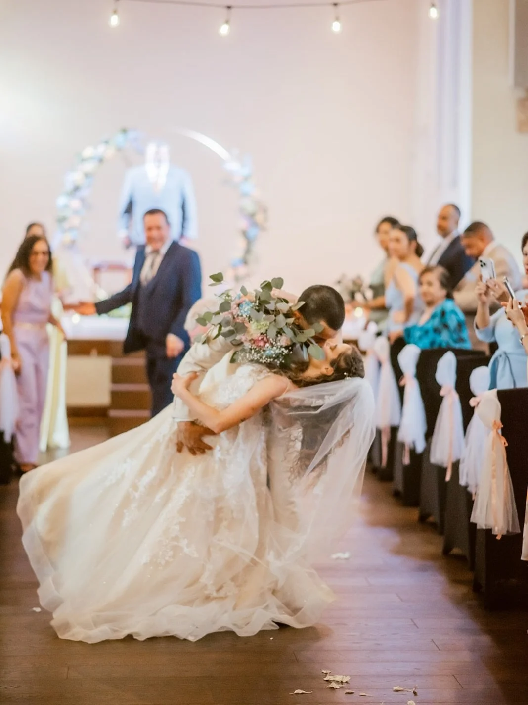 A bride and groom kiss during their wedding ceremony, with guests watching and taking photos. The bride holds a bouquet of flowers, and the groom wears a suit. The setting is decorated with flowers and string lights.