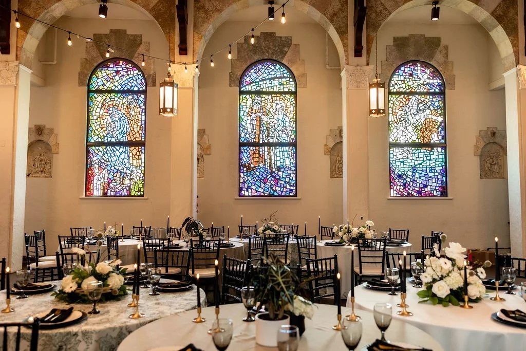 Elegant banquet hall with round tables decorated with white flowers, black chairs, and candles at each table. There are three stained glass windows with colorful mosaic designs on the back wall, and string lights hanging from the ceiling. The room has high arches and decorative wall niches.