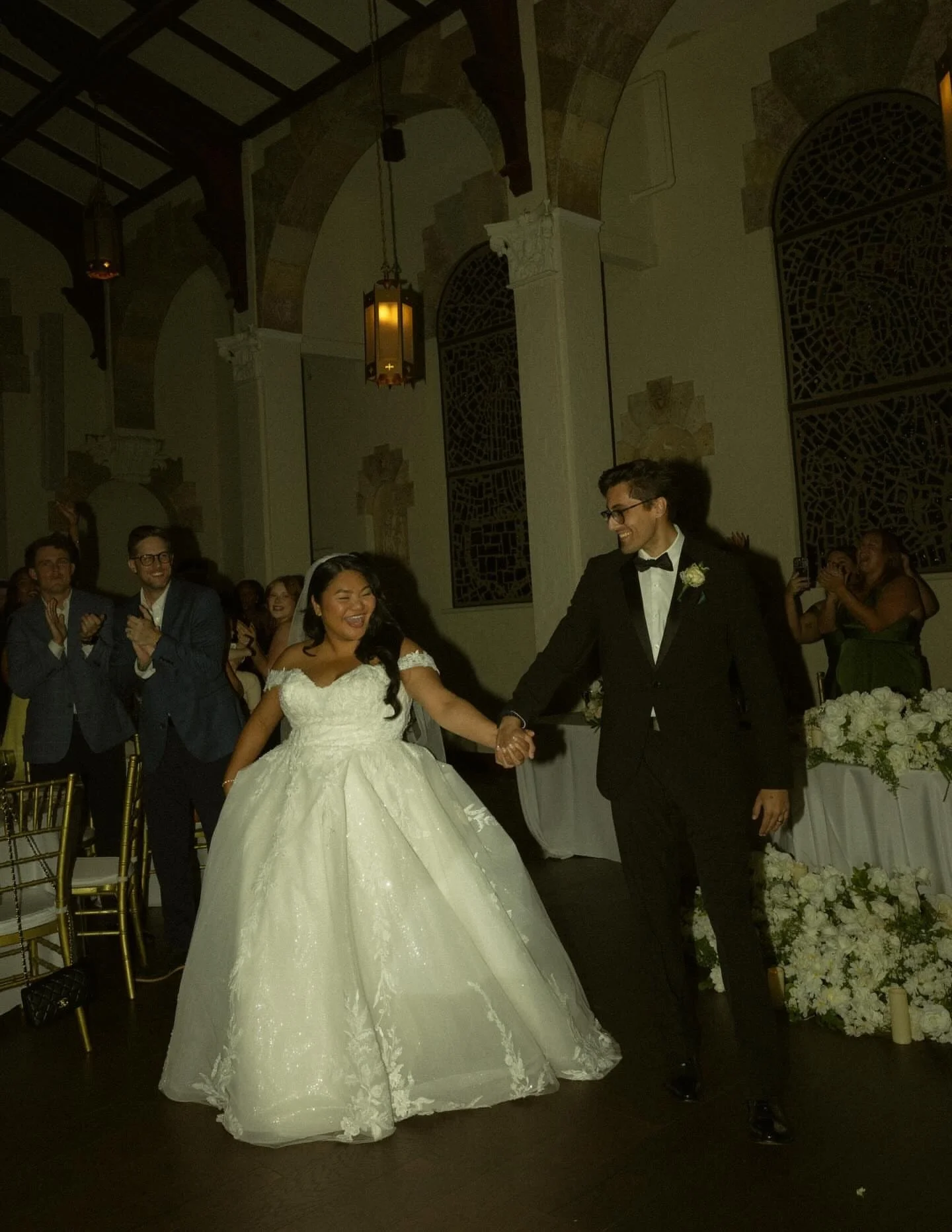 A bride and groom holding hands and smiling during their wedding reception, with guests clapping and taking photos in a decorated hall.