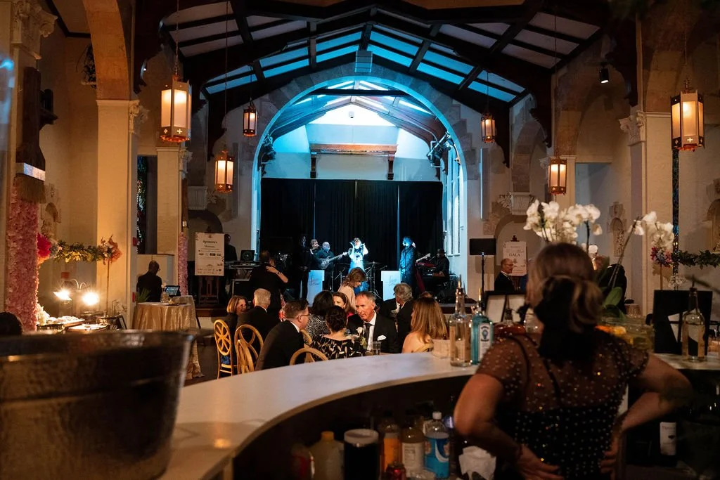 Guests dining and socializing at tables in a large, elegant event hall with a stage where a band is performing, and floral decorations on the tables.