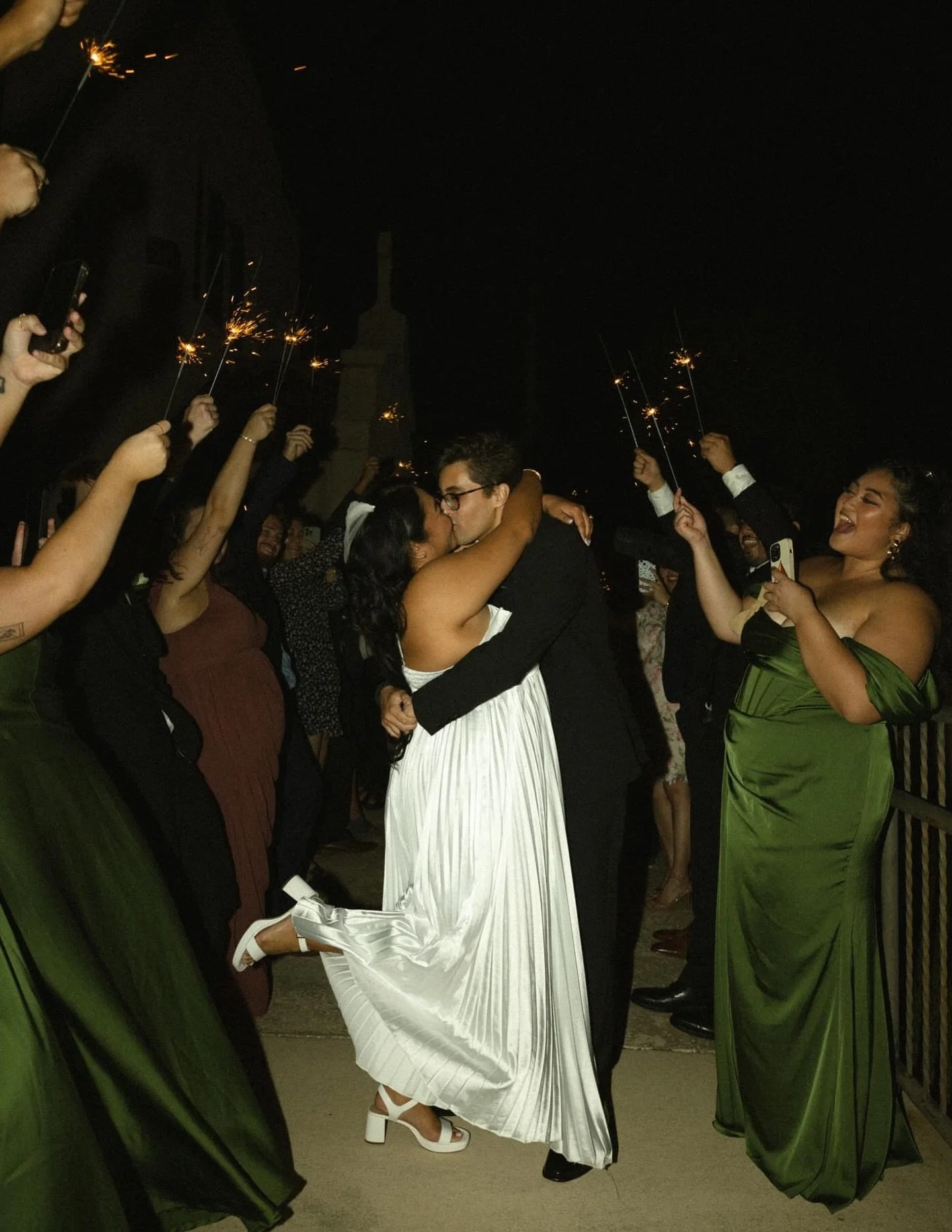 A couple is kissing while dancing at night surrounded by friends holding sparklers, celebrating at a festive event.