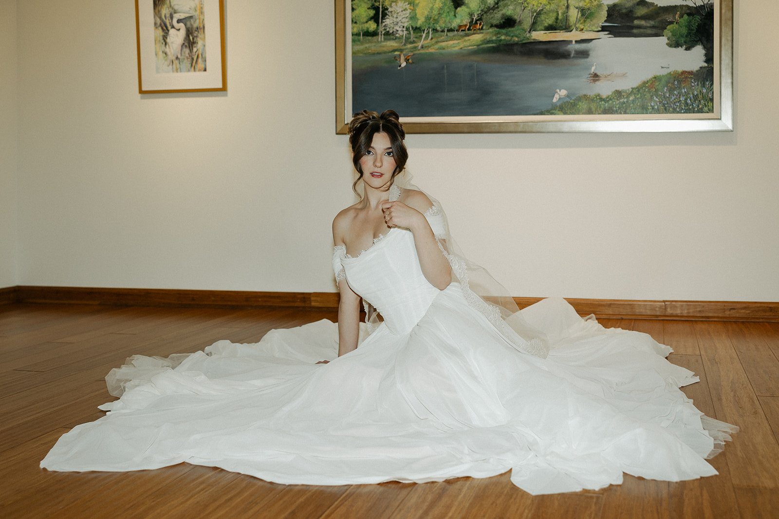 A woman in a wedding dress sitting on a wooden floor in front of a plain wall with two paintings, one of a lake with trees and birds, and the other of a bird.