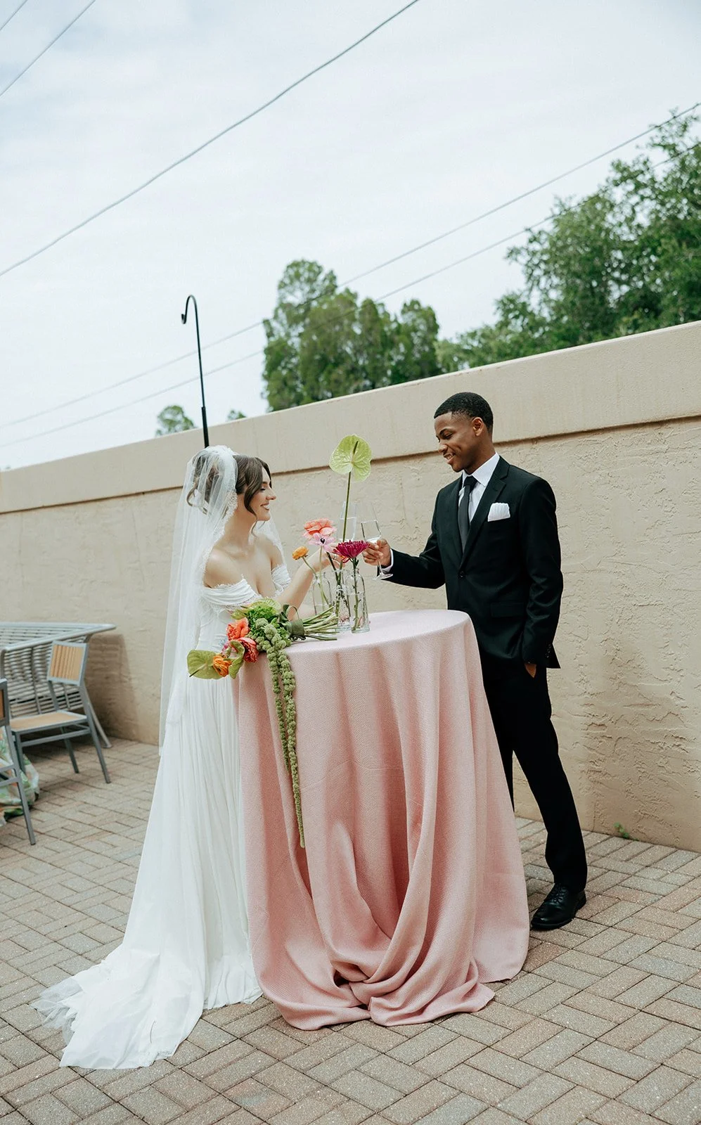 Bride and groom in wedding attire at an outdoor celebration, toasting with wine glasses over a table with flowers.
