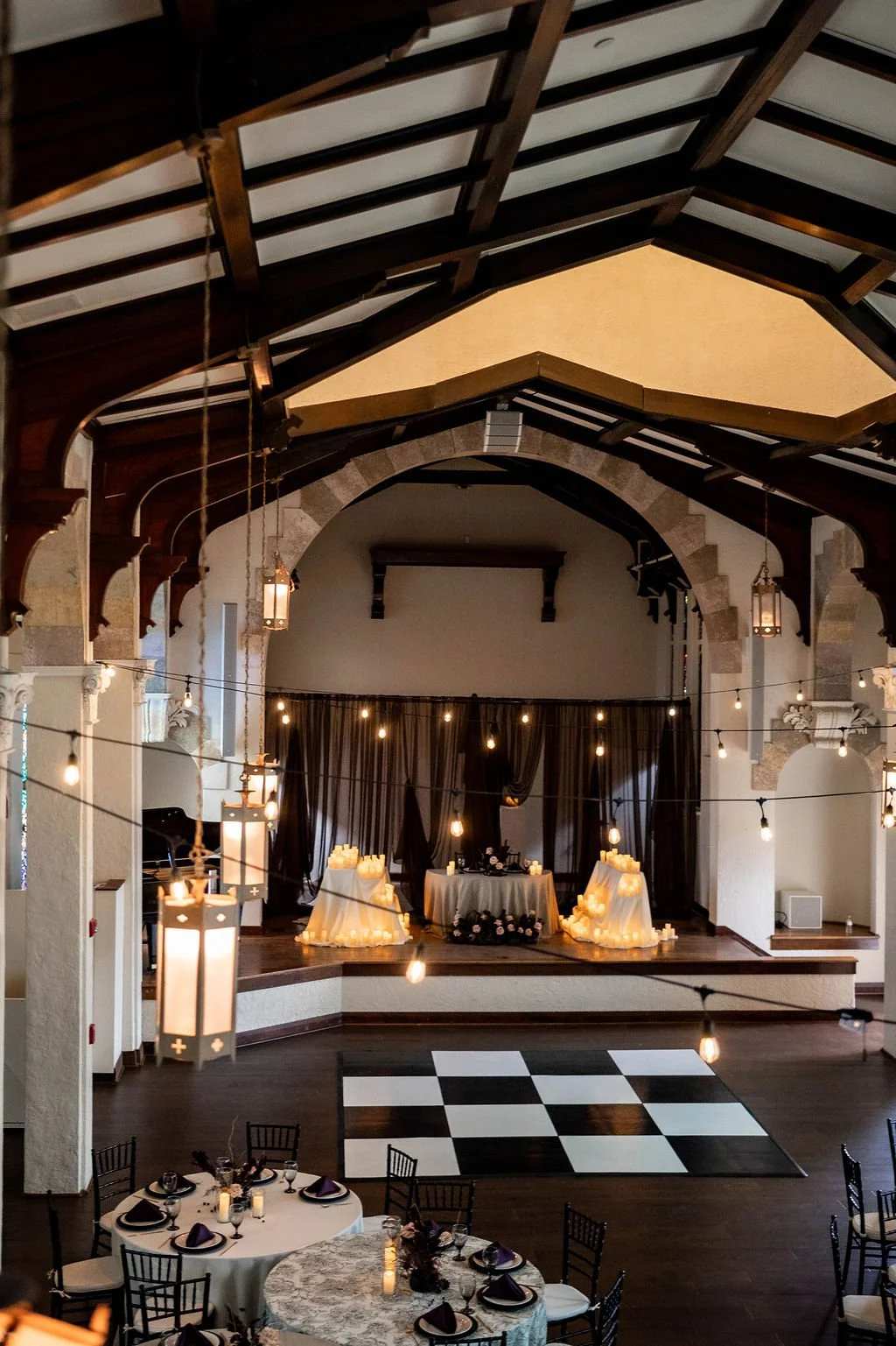Elegant event space with a small stage decorated with candles, drapery, and flowers, surrounded by string lights, with dining tables set for guests.