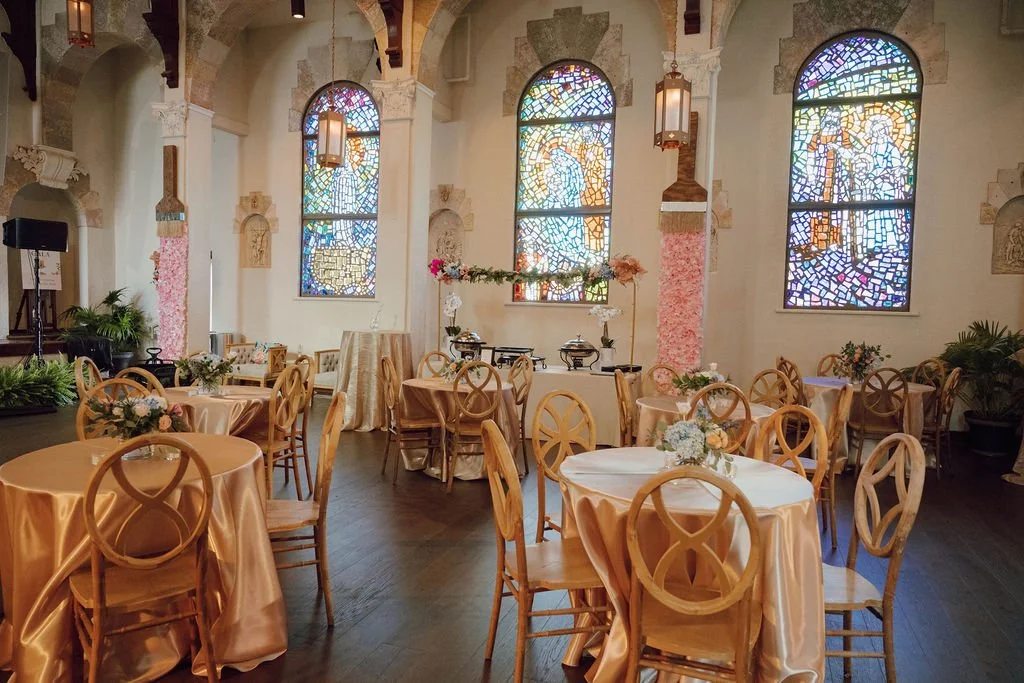Elegant event hall with round tables covered in peach-colored tablecloths, decorated with floral centerpieces, set in front of three stained glass windows, with pink and white flowers on some tables, and a small buffet setup on the back wall.