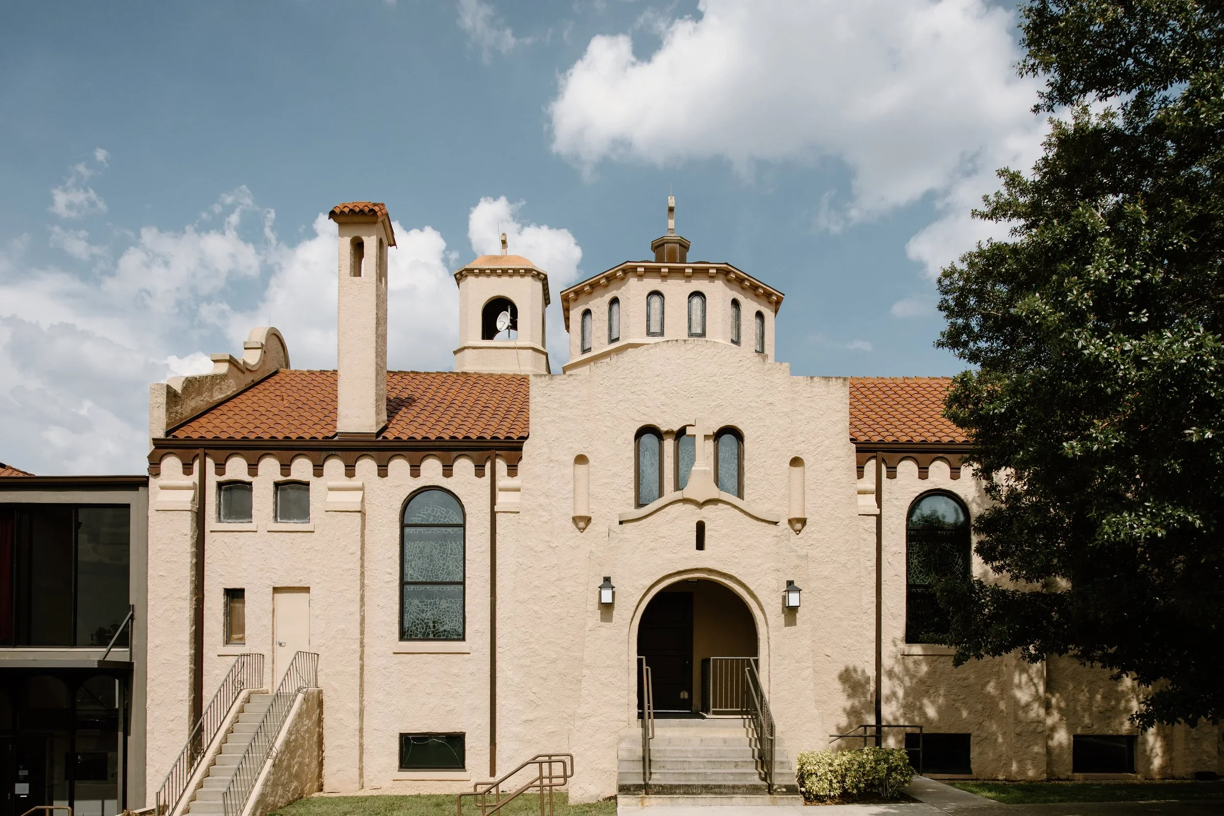 A beige stucco building with red tile roof, arched door, and multiple small towers, surrounded by greenery and a partly cloudy sky