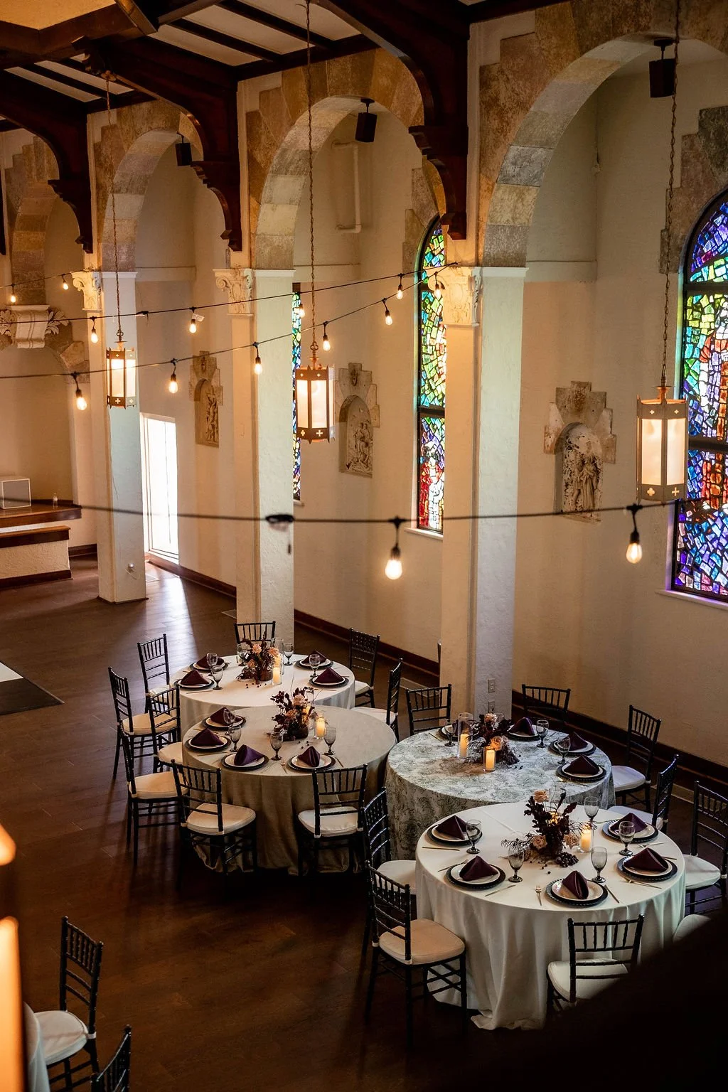Wedding reception setup in a church with round tables decorated with fall-themed centerpieces, surrounded by black chairs, lit by hanging string lights and chandeliers, featuring arched stained glass windows.