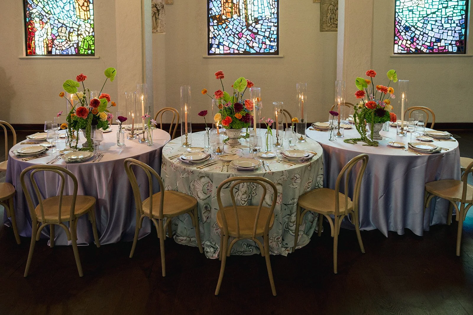 Three round banquet tables set for a formal event with floral centerpieces and candlelit tall candlesticks in an elegant room with stained glass windows.