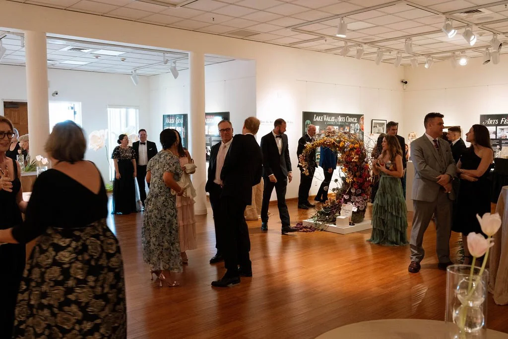 People in formal attire gather at an indoor art gallery event, with floral art installations and information boards showcasing local arts and community organizations.