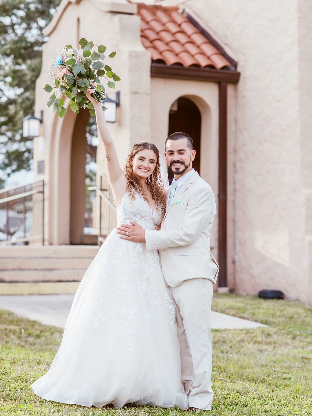 A happy couple in wedding attire posing outdoors in front of a building, with the woman holding a bouquet of greenery and flowers raised in the air.