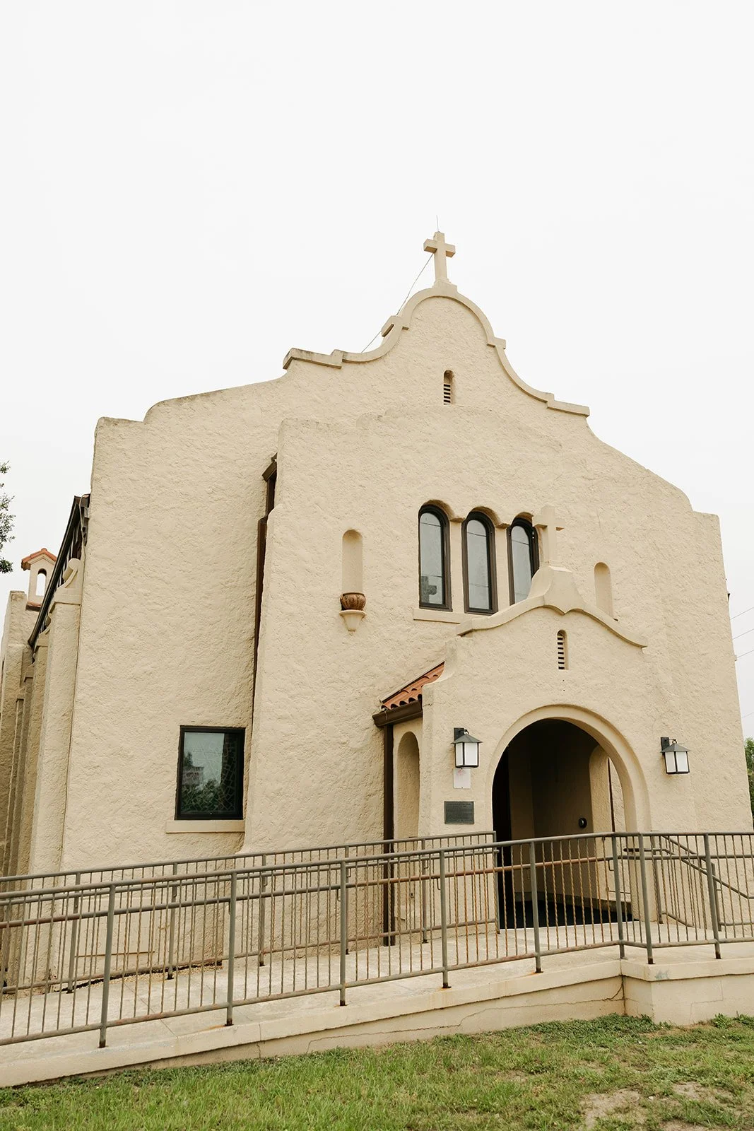 A beige stucco church with a cross on top, arched windows, and an arched entrance, surrounded by a metal railing and a grassy area.