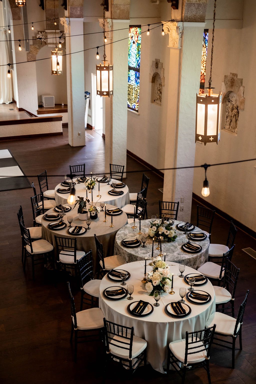 Elegant event space with round tables set for a formal dinner, decorated with floral centerpieces, candles, and black plates and napkins at Gather at Lake Wales Art Center