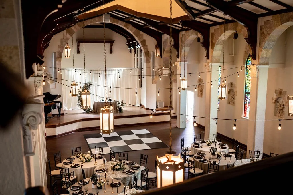 Elegant event space with round tables set for a formal dinner, decorated with white and black floral centerpieces, in a large hall with high arched ceilings, hanging lights, stained glass windows, and a stage at the front.