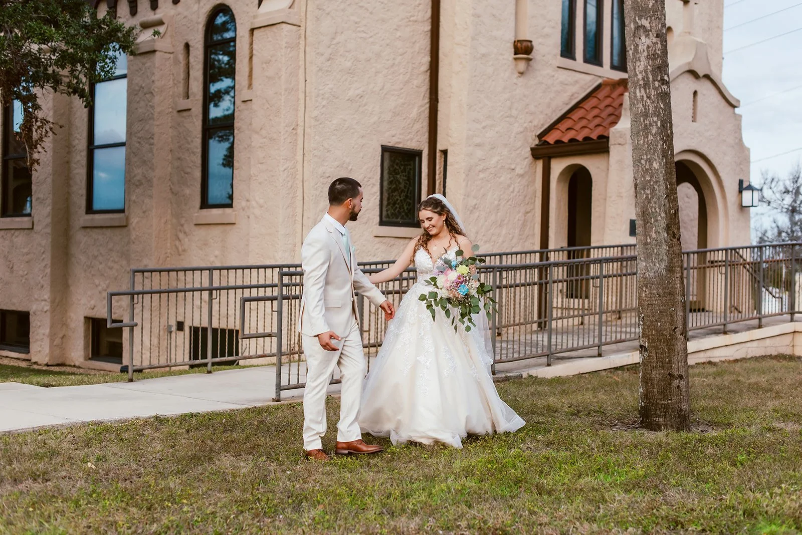 A bride and groom smiling and holding hands outside a building, with the bride holding a colorful bouquet, during their wedding.
