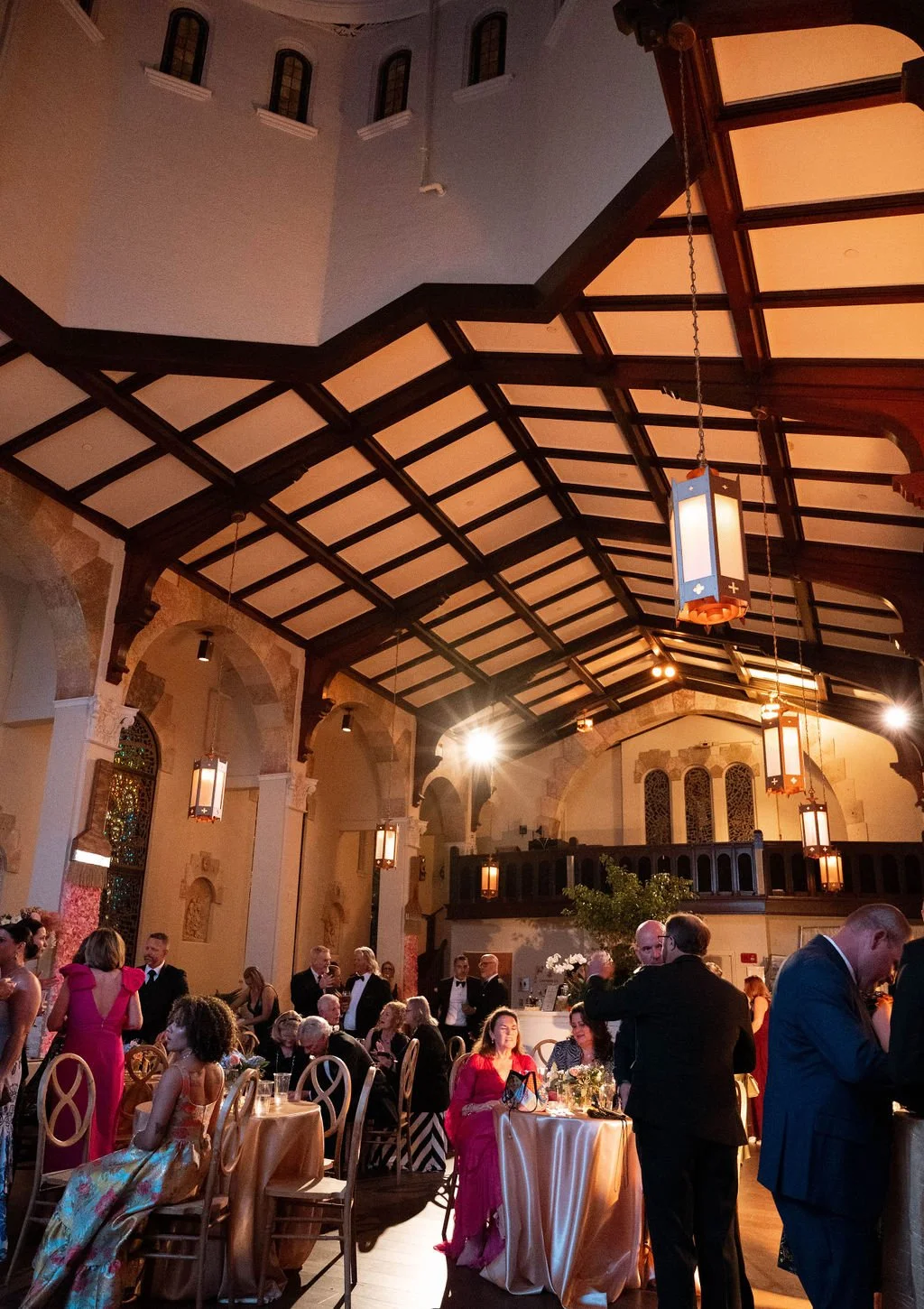 People gathering at a formal event inside a church-like venue with high vaulted ceilings, hanging lamps, and arched windows, some sitting and others standing and talking.