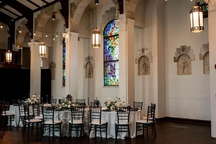 Round tables with white tablecloths, floral centerpieces, and black chairs set up in a church with stained glass windows and hanging lanterns.