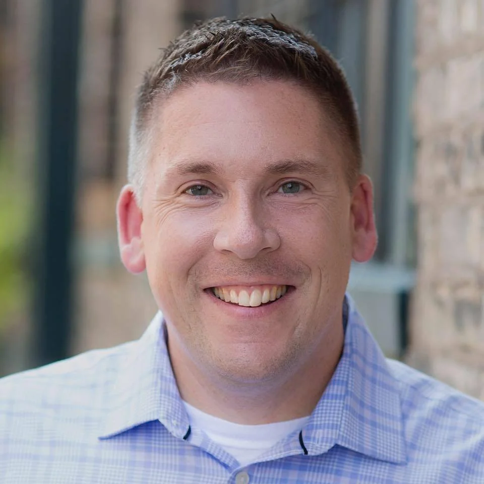 Close-up of a smiling man with short brown hair, wearing a light blue checkered shirt, outside near a brick wall.